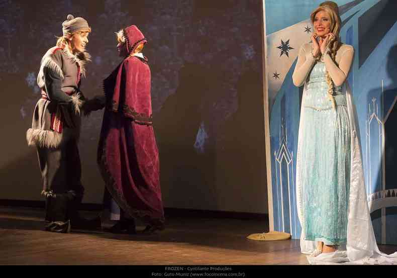 Dois atores esto do lado esquerdo do palco, enquanto atriz loura, com a roupa azul de Frozen, sorri,  esquerda 