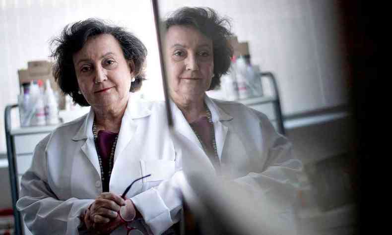 Médica e pesquisadora Margareth Dalcomo, usando jaleco e de braços cruzados, olha para a câmera e tem imagem refletida no espelho 