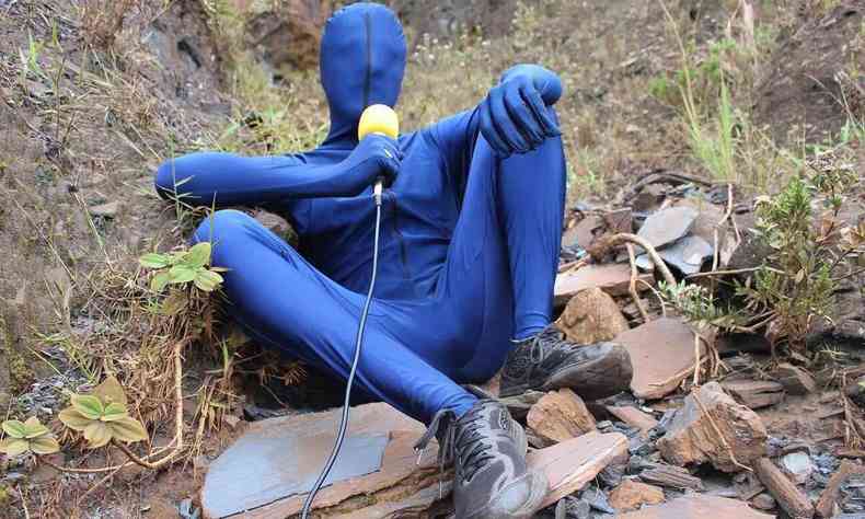 Homem vestido com fantasia azul, que cobre seu rosto e corpo, segura microfone amarelo no curta Vida terrena no identificada 