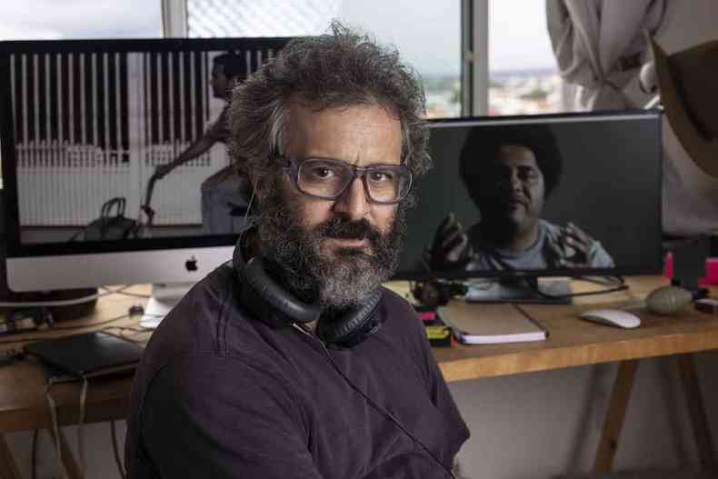 Cineasta Adirley Queirós está sentado, tendo ao fundo a tela do computador com a cena de um filme 