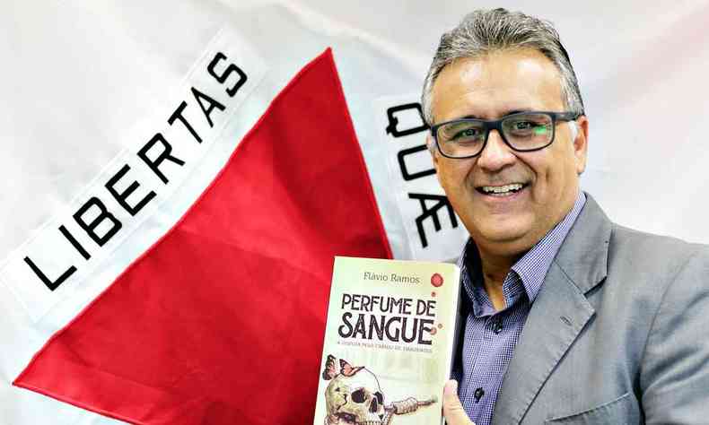 O autor Flvio Ramos segura seu livro 'perfume de sangue', tendo ao fundo a bandeira de Minas Gerais