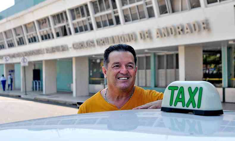 Taxista de blusa laranja que atua nas imediaes do Aeroporto da Pampulha em p, prximo ao seu veculo, que  branco e carrega uma sinalizao de txi