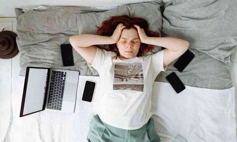 Mulher deitada na cama com expressão angustiada, rodeada por um computador e celulares