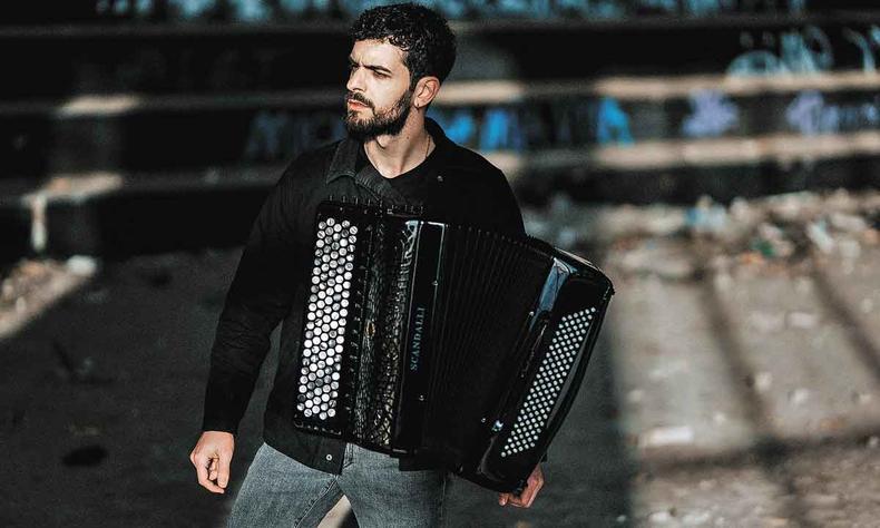de cala jeans e camisa preta, com um acordeom, o msico Pietro Roffi olha para ponto fora de quadro