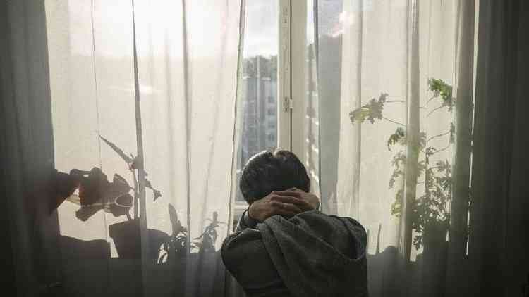 Homem com cabelos pretos  visto de costas, com as mos na nuca, olhando por uma janela