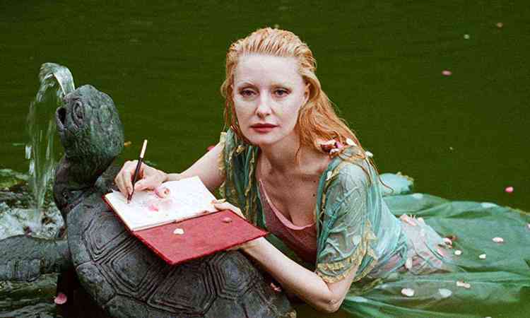 Mulher loura e bonita escreve dentro de um rio, deitada, usando vestido verde e com livro apoiado na cabea da escultura de um sapo