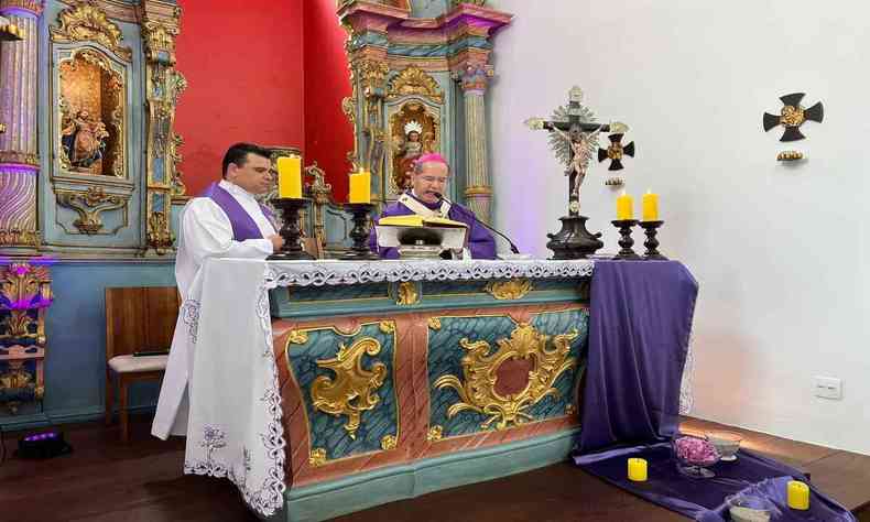 Um padre em cima do altar celebrando a missa. Sua roupa  branca e de cor roxa. Muitos cruzes e um arquitetura colonial com muitos detalhes podem ser vista no altar