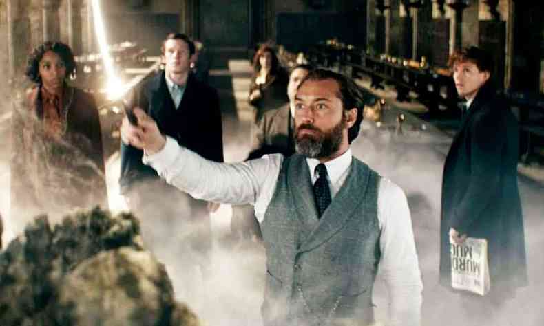 de terno cinza, camisa branca e braos cruzados, Jude Law, cercado por outros atores de animais fantsticos, olha para cima, em cena de o segredo de dumbledore