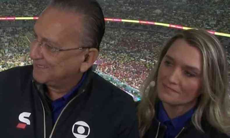 Imagem em close nos rostos de Galvo Bueno, um homem branco de cabelos rasos e grisalhos que usa um culos de grau e camiseta azul com o logo da Globo. e Ana Thas Monteiro, uma mulher branca e loira de olhos claros com a mesa camisa de Galvo. Ele d as costas  comentarista, que olha em direo a ele