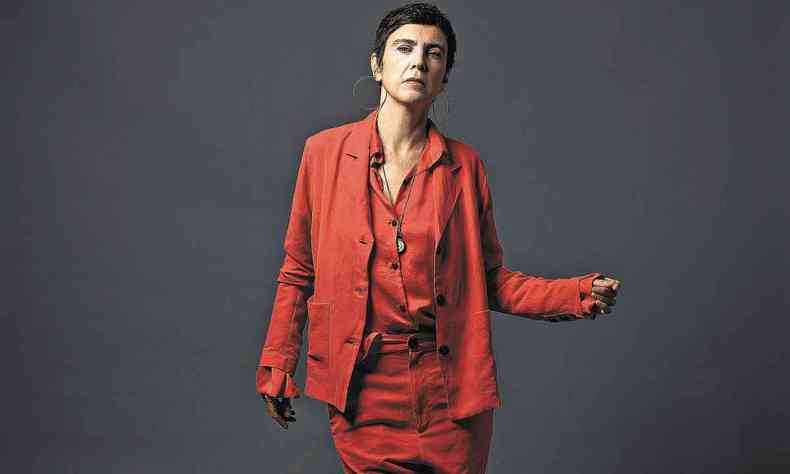 De cala, camisa e palet vermelhos, Adriana Calcanhotto posa para foto, de p, com fundo neutro