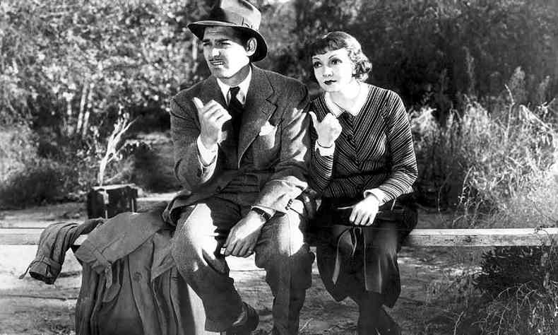 Os atores Clark Gable e Claudette Colbert, sentados num banco rstico de madeira, com o mato em volta deles, fazem o gesto de carona no filme Aconteceu naquela noite