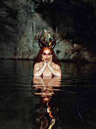 maquiada e com adereos pontiagudos na cabea, Gloria Groove aparece com o corpo parcialmente mergulhado em lago dentro de gruta, segurando o queixo com as mos 