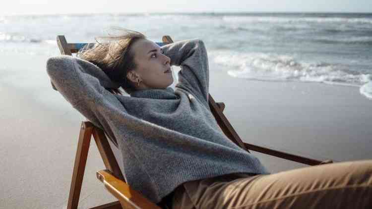 Mulher deitada em cadeira em praia, com os braços atrás da cabeça e olhando para o 'nada'