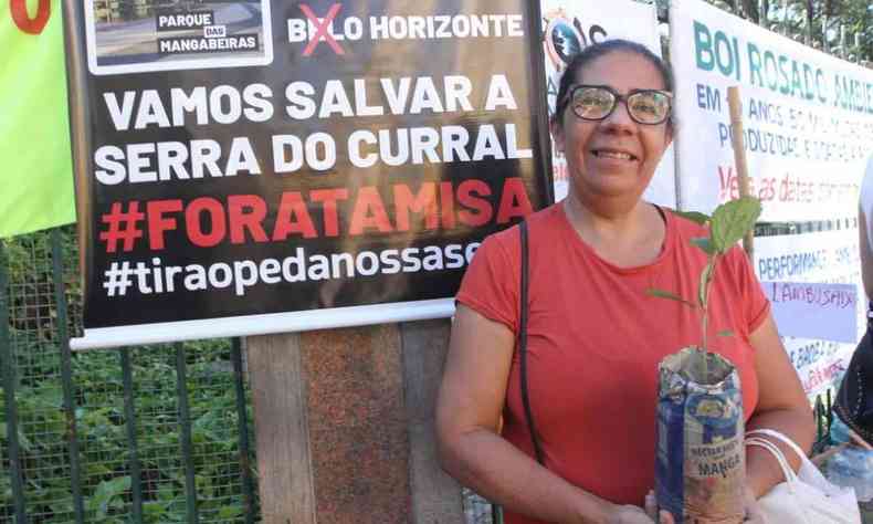 Juliana Maria Ferraz usa camisa vermelha e segura na mo um muda de ip branco, atrs dela um cartaz com os dizeres 'Vamos salvar a Serra do Curral. Fora Tamisa'