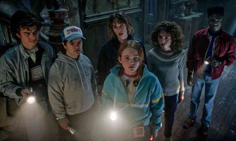 Seis garotos, tendo à frente jovem segurando lanterna acesa, olham preocupados para o alto, na penumbra, em cena de Stranger things