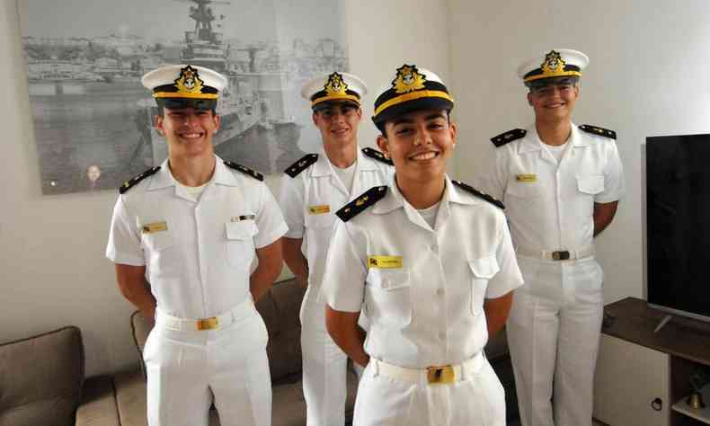 Quatro jovens fardados com uniformes da Marinha Brasileira posam em frente a um quadro de navio