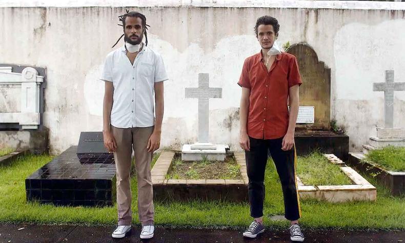 Dois rapazes usando cala, camisa, tnis e mscara, de p em frente a uma sepultura, encaram a cmera em cena de garotos ingleses