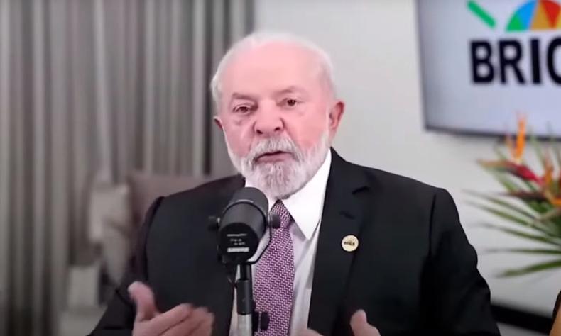 Lula, usando terno preto e gravata listrada bord, falando ao microfone. ao fundo a logo do Brics