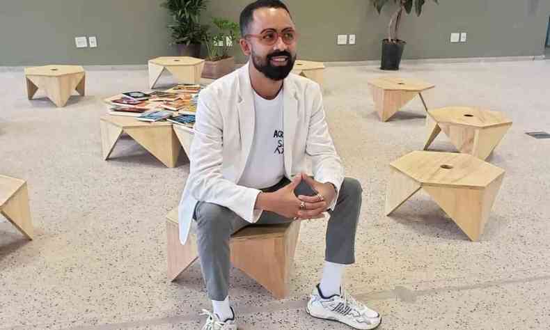Artista Jaime Lauriano sentado em meio a banquinhos com capas de livros de autores negros na biblioteca de Inhotim