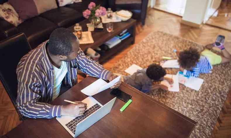 Pai trabalhando  mesa enquanto filhos desenham no tapete ao lado