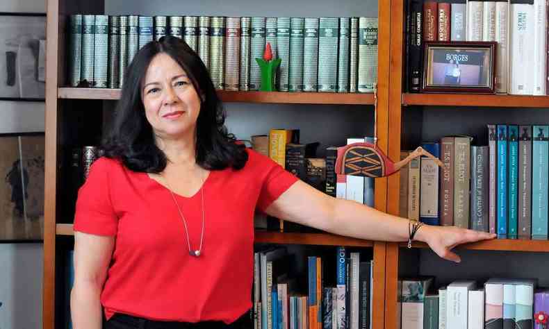 A escritora e professora Maria Esther Maciel sorri e tem a mo direita na prateleira de estante repleta de livros