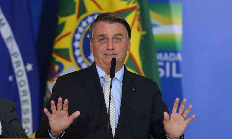 O presidente Jair Bolsonaro de palet preto, blusa azul-claro e gravata amarela com listras azuis. Com expresso sorridente, ele fala ao microfone e mostra as palmas das mos. Ao fundo, a bandeira do Brasil.