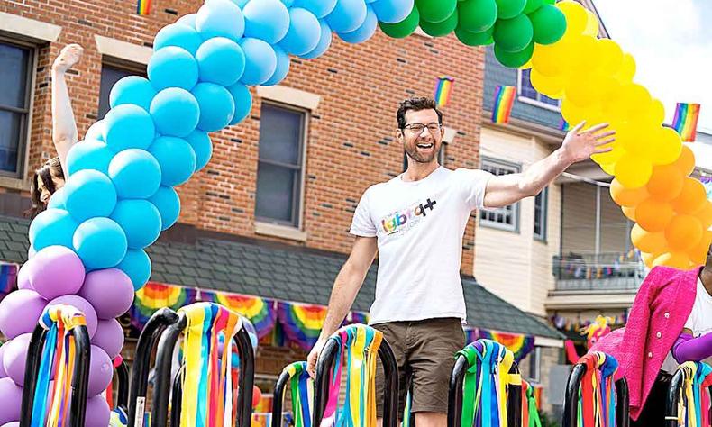 Com camisa escrita LGBTQ+, ator ergue o brao e sorri, debaixo de arco-iris colorido feito com bales, em cena de mais que amigos 