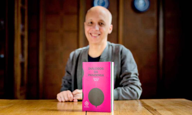 Livro 'Dirios da pandemia' est numa mesa. Ao fundo, sentado, est o jornalista Helvcio Carlos, coordenador da edio