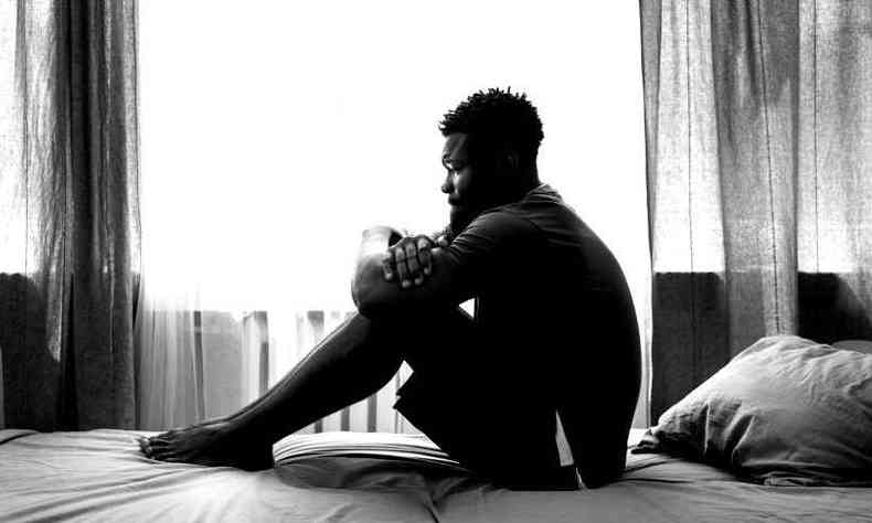 Homem negro sentado em sua cama enquanto abraa as prprias pernas. A imagem est em preto e branco. Atrs dele, h uma cortina.