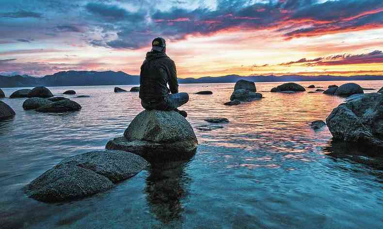 Homem pratica meditao, sentado numa pedra dentro do mar, de frente para o nascer do sol