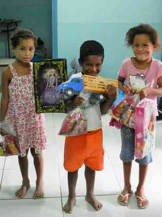Duas meninas e um menino seguram brinquedos e alimentos doados