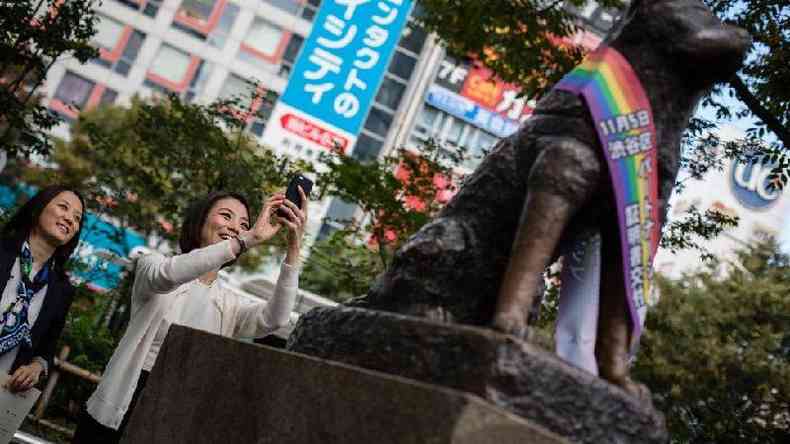 Mulher tirando foto da esttua de Hachiko que est com uma faixa com as cores do arco-ris