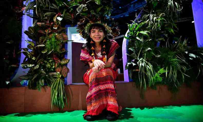 A líder indígena brasileira Txai Suruí sorri, usando cocar e roupa típica de sua tribo, sentada num palco durante a COP-26, na Escócia