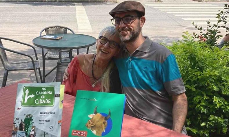Escritora Ana Raquel est sentada em mesa, na rua, com o rosto no ombro do filho Daniel Barcellos. Na frente deles esto livros lanados pela dupla 