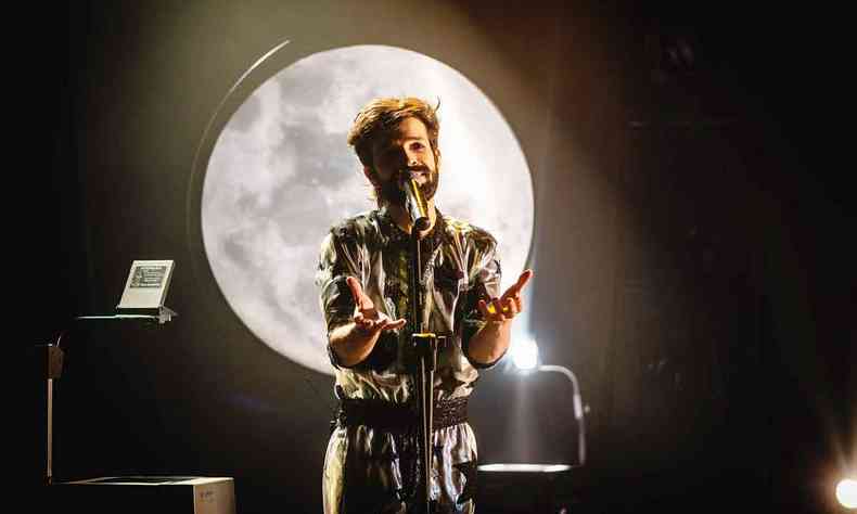 Cantor Felipe de Oliveira estende os braos, em volta do microfone, e tem a lua no fundo do cenrio de seu show
