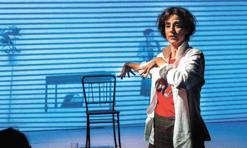A atriz Mariana Lima gesticula com as mãos, tendo ao fundo tela azul e uma cadeira, em cena da peça ''Cérebro_Coração''