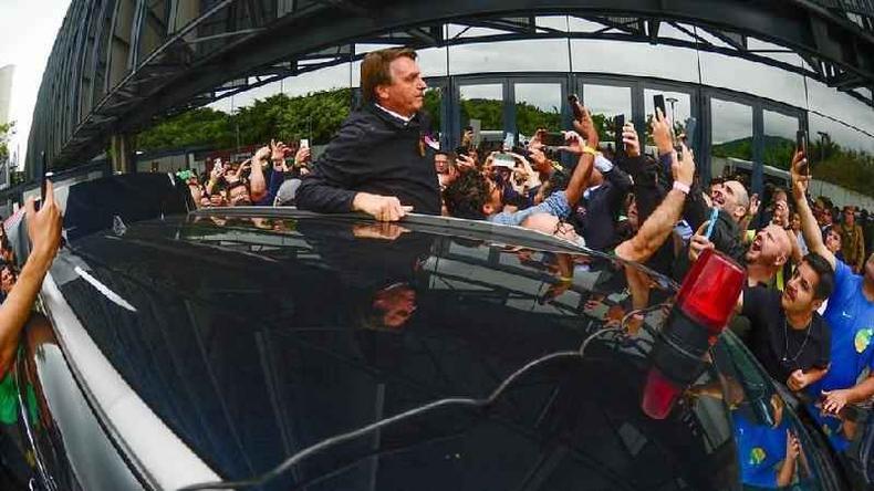 Bolsonaro em p para fora do carro, enquanto pessoas tentam tirar foto com ele