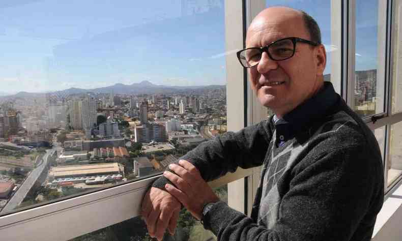 Escritor Luiz Ruffato olha para a câmera, diante de janela com a ensolarada Belo Horizonte ao fundo