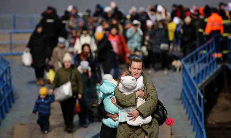 Fila de pessoas refugiadas.  frente, uma mulher branca de cabelos castanhos claros com uma criana no colo.