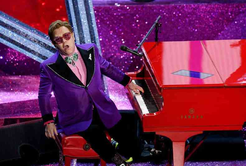 Elton John, de terno roxo e óculos escuros, toca um piano vermelho no palco do Oscar 2020