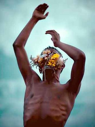 Rapaz com torno nu e arranjo de flores cobrindo o rosto estica os braos em direo ao cu