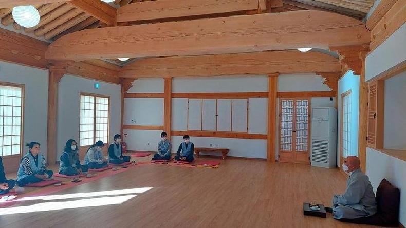 Um grupo de homens e mulheres sentados de pernas cruzadas na frente de um monge em uma sala escassamente decorada em um templo budista