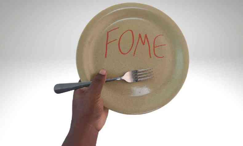 A foto tem o fundo brando e mostra a mo de uma pessoa negra segurando um garfo e um prato de cermica bege onde est escrito 'fome' em vermelho