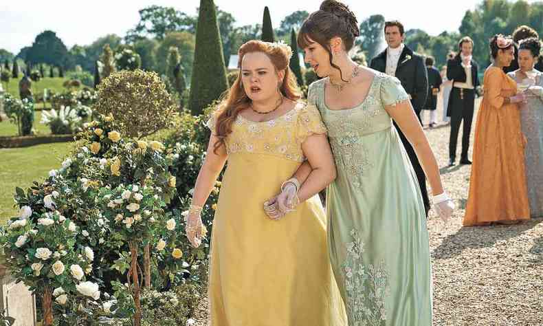 As atrizes Nicola Coughlan e Claudia Jessie, vestidas com longos de festa e luvas caminham de mos dadas por jardim, com semblantes preocupados em cena de Bridgerton