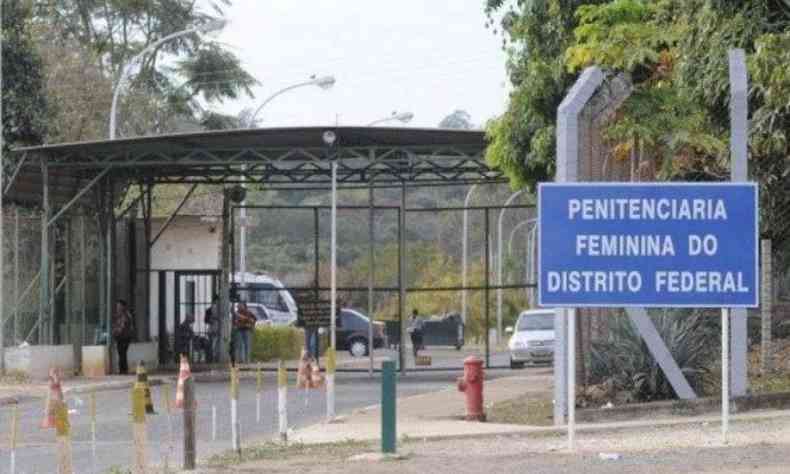 Imagem externa da Penitenciria Feminina do Distrito Federal. H uma placa azul  direita com o nome do local e, ao fundo e ao centro, uma grade que se liga a uma portaria.