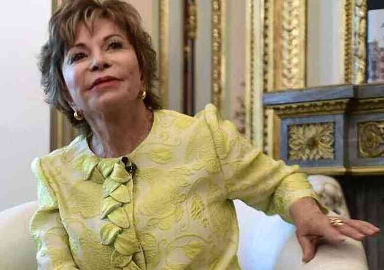Isabel Allende sentada numa cadeira branca, vestindo calas pretas e casaco amarelo dourado, com brincos e anis de ouro