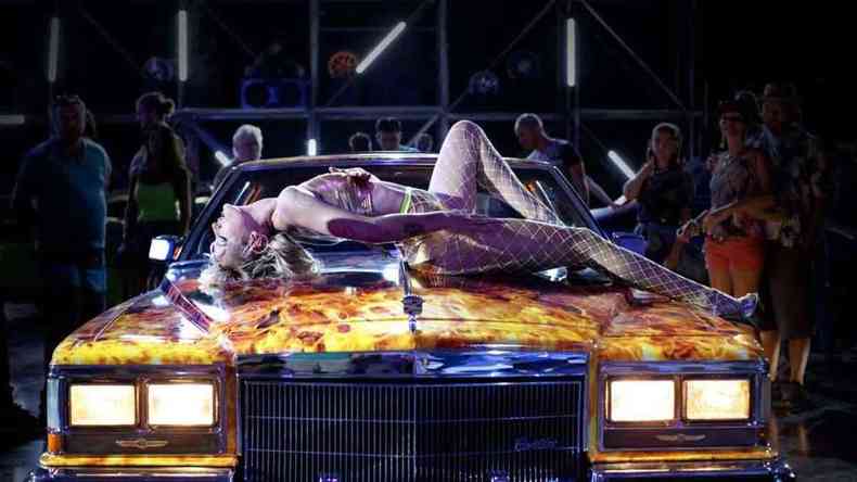Pessoas observam mulher de biquni deitada sensualmente sobre um carro