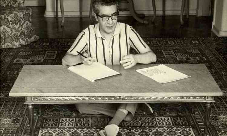 Escritor Campos de Carvalho est sentado no cho, sobre o tapete, escrevendo sobre a mesa de centro de uma sala
