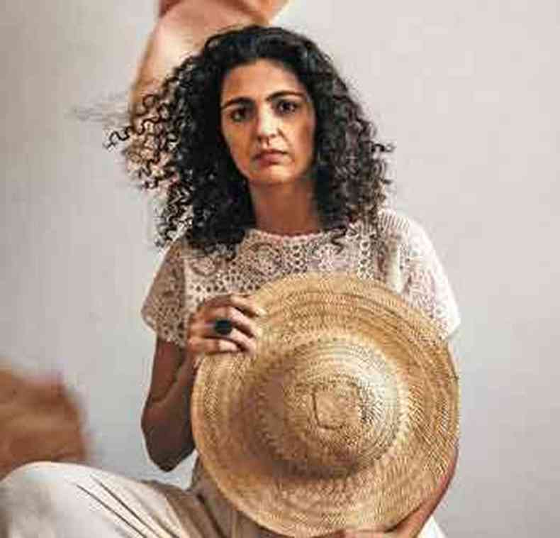 de cala e blusa brancas, segurando chapu de palha, Luiza Brina sentada num banco numa sala com obra de arte na parede
