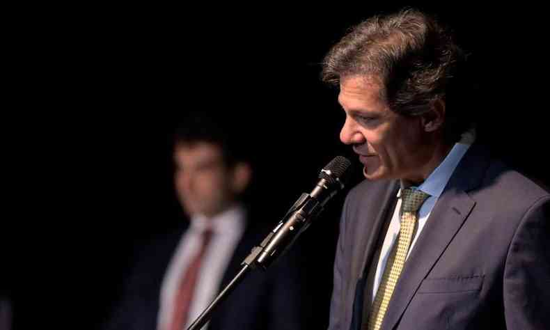 A imagem mostra Haddad, no canto direito da foto, falando ao microfone. Ele  um homem branco de cabelos lisos e preto e usa terno